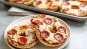 Close-up of four golden mini pizza bagels topped with melted cheese, pepperoni slices, and dried herbs, served on a beige plate.