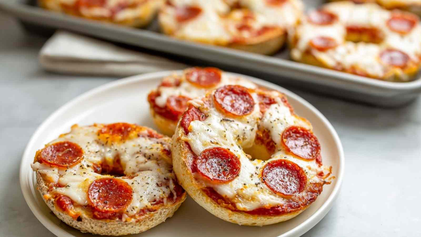 Close-up of four golden mini pizza bagels topped with melted cheese, pepperoni slices, and dried herbs, served on a beige plate.