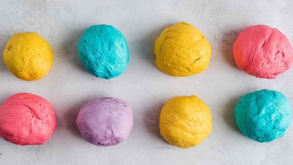 Eight vibrant dough balls in red, yellow, purple, teal, and pink, ready for shaping into rainbow bagels.