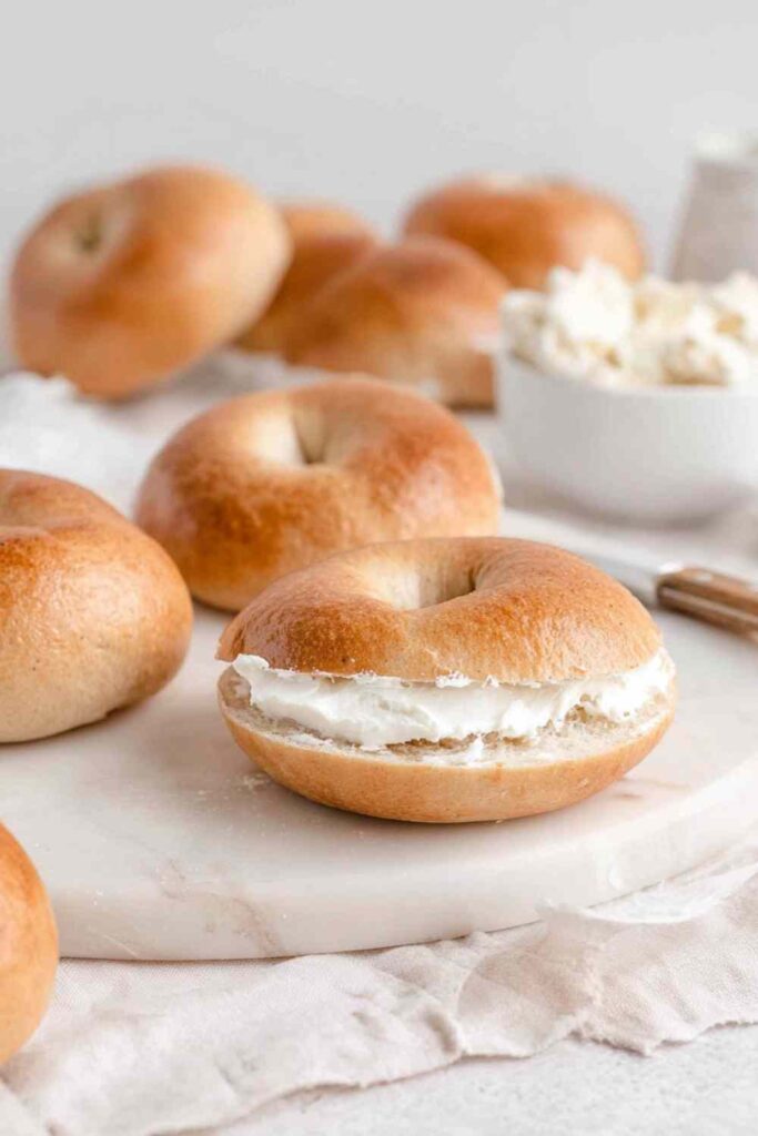 Close-up of a mini bagel sliced and filled with cream cheese, surrounded by other bagels and a bowl of cream cheese in the background.