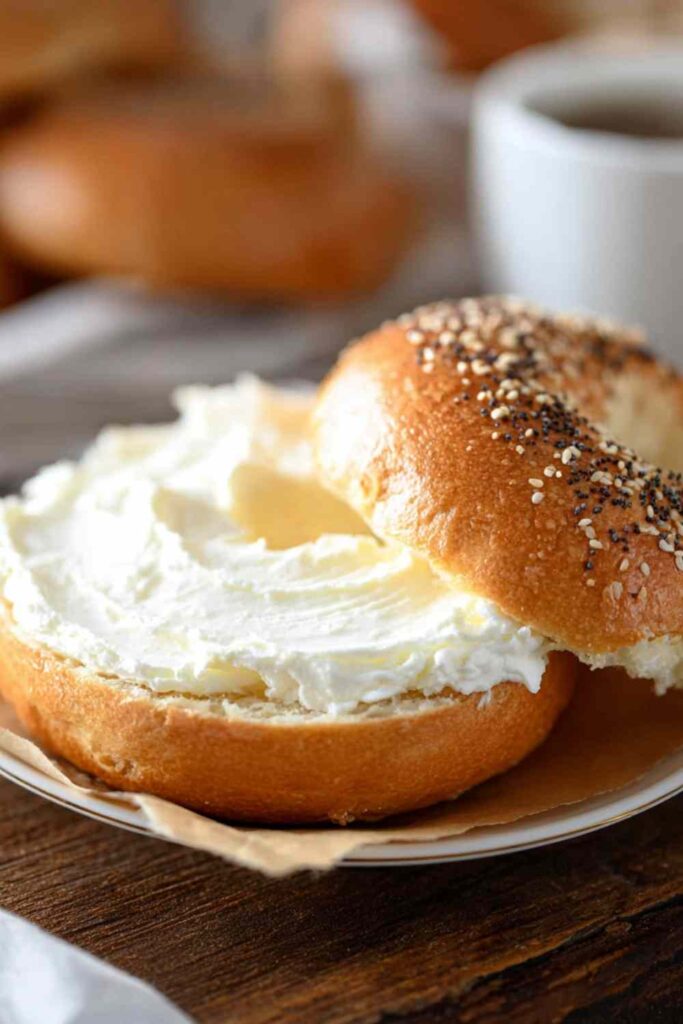 Sliced gluten free bagel topped with thick cream cheese, sprinkled with seeds, served with coffee in background