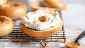 Open New York-style bagel generously spread with whipped cream cheese, resting on a cooling rack with seasoning dusted nearby