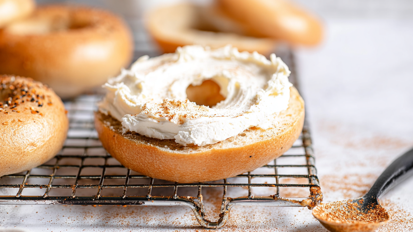 Open New York-style bagel generously spread with whipped cream cheese, resting on a cooling rack with seasoning dusted nearby