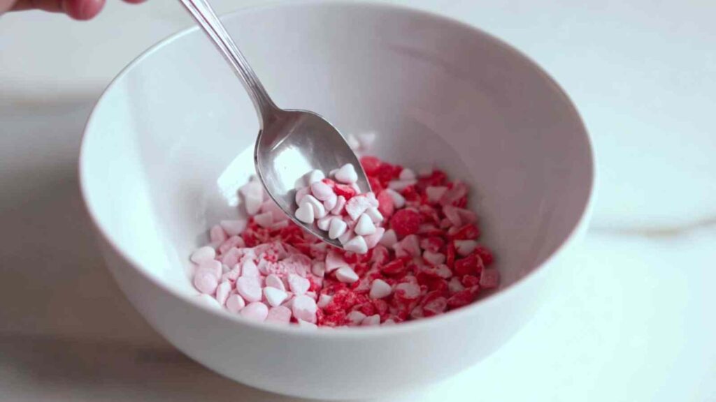 Spoon scooping crushed pink and white candy hearts from a white bowl—ideal for sweet bagel toppings.