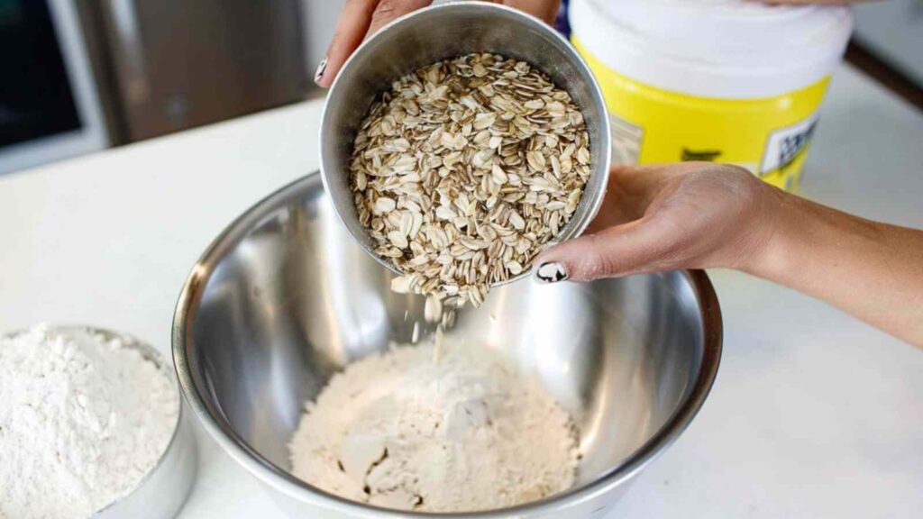 Hand pouring rolled oats into a stainless bowl of oat flour, preparing dry ingredients