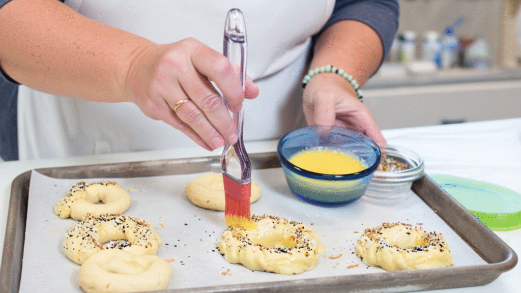 Person brushing egg wash onto raw low carb bagels topped with seasoning on a baking tray