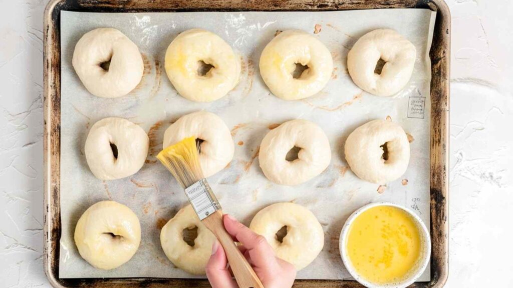 Hand brushing egg wash over unbaked mini bagels on a parchment-lined baking sheet with a bowl of egg wash nearby.