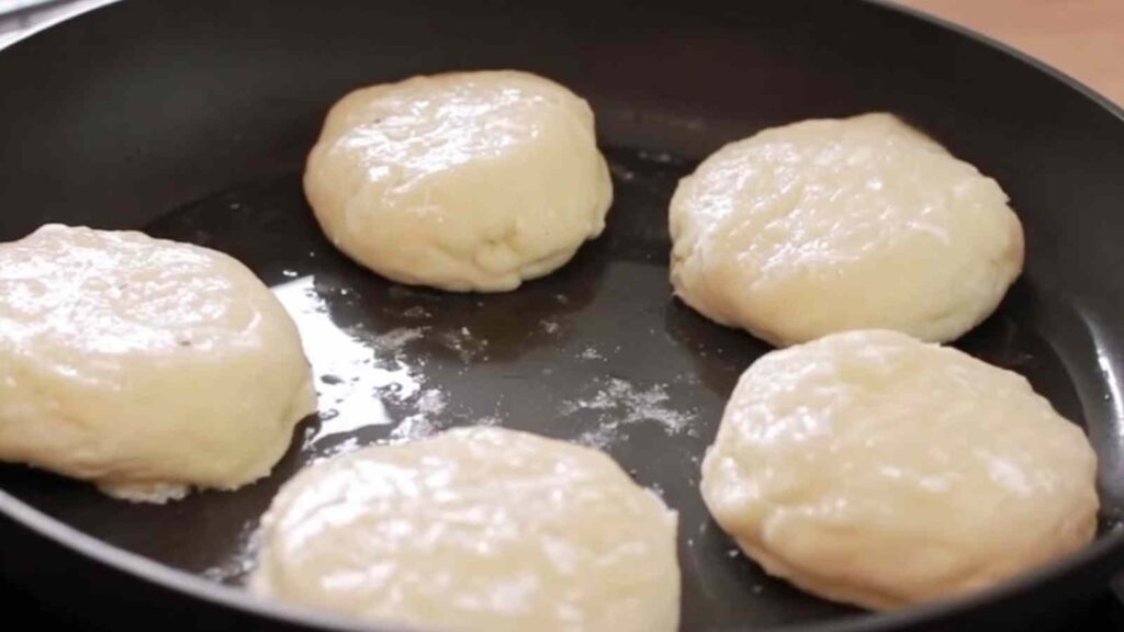Raw English muffin rounds cooking in a nonstick skillet, rising gently as they steam under a lid