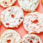Overhead view of open bagels topped with a variety of cream cheese spreads on a coral background.