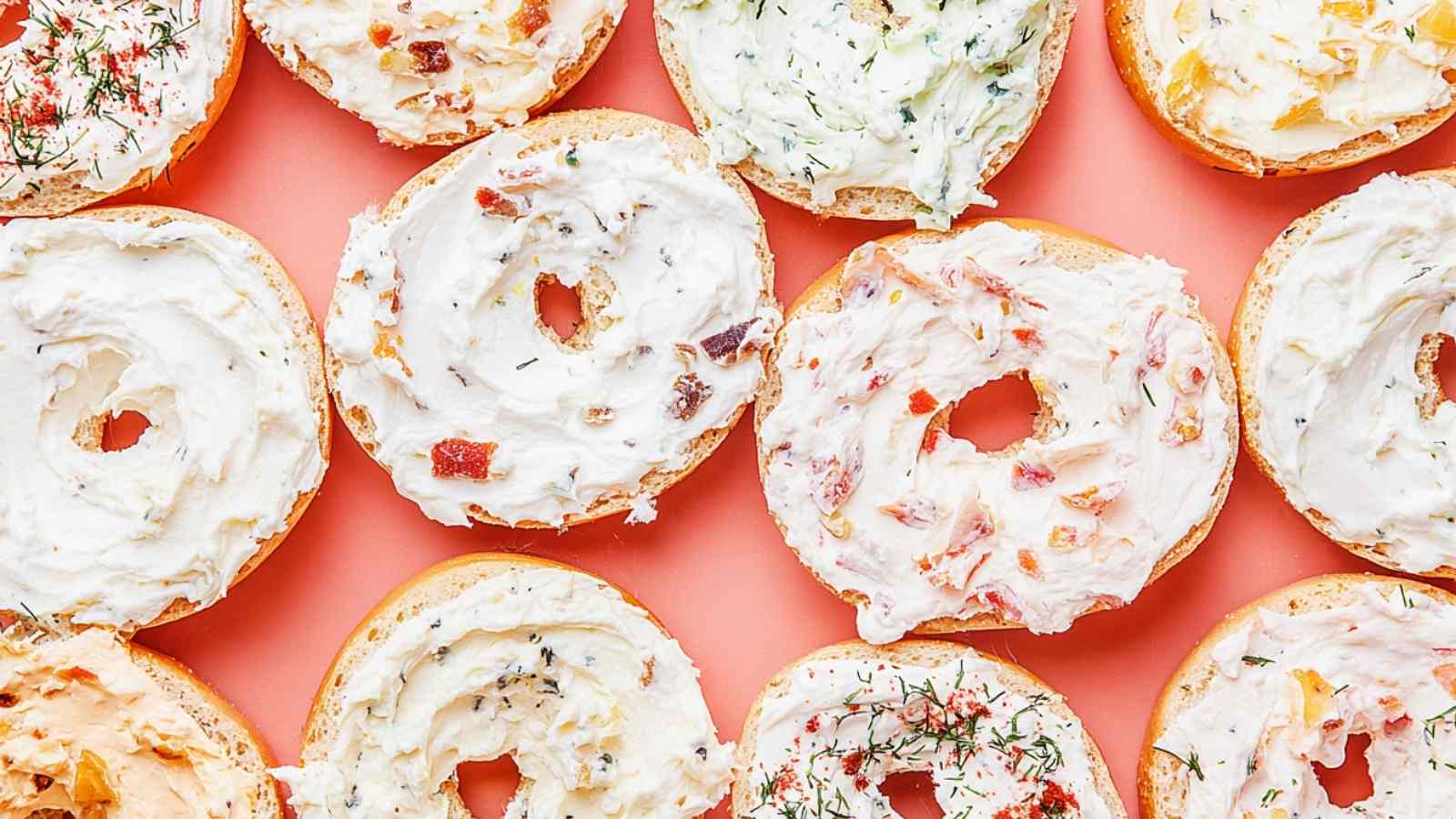 Overhead view of open bagels topped with a variety of cream cheese spreads on a coral background.