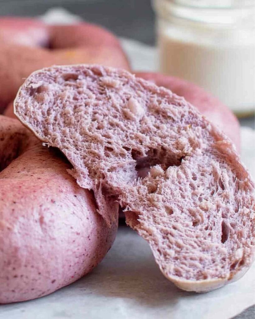 Close-up of a sliced strawberry bagel showing its airy, fluffy interior with a vibrant natural pink hue, set against whole pink bagels.

