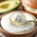 Spoon dipping into a bowl of whipped cream cheese with bagel and avocado in background