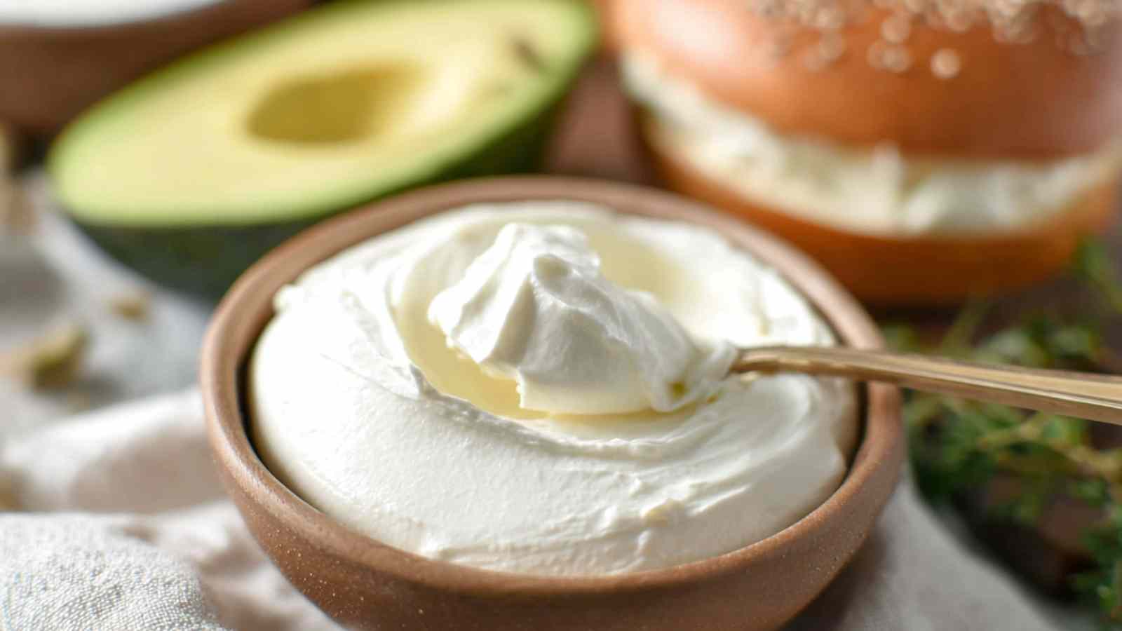 Spoon dipping into a bowl of whipped cream cheese with bagel and avocado in background