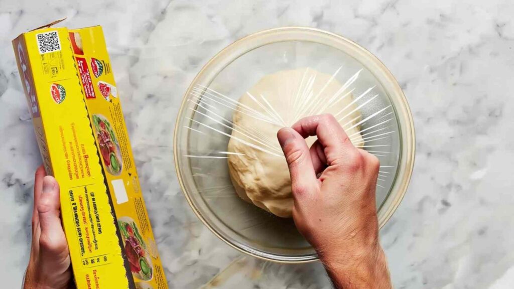 Bowl of bagel dough covered with plastic wrap, hand finishing the seal