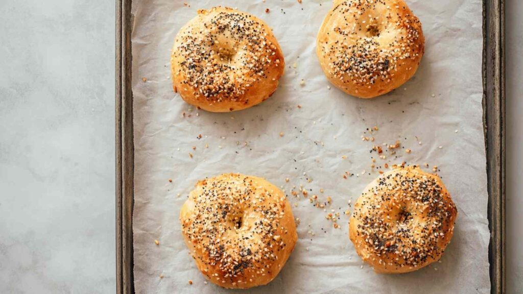 Four freshly baked golden-brown bagels with everything seasoning on a parchment-lined baking tray.