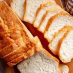 Overhead view of a golden loaf of sliced protein bread on wooden board with light crust and soft crumb