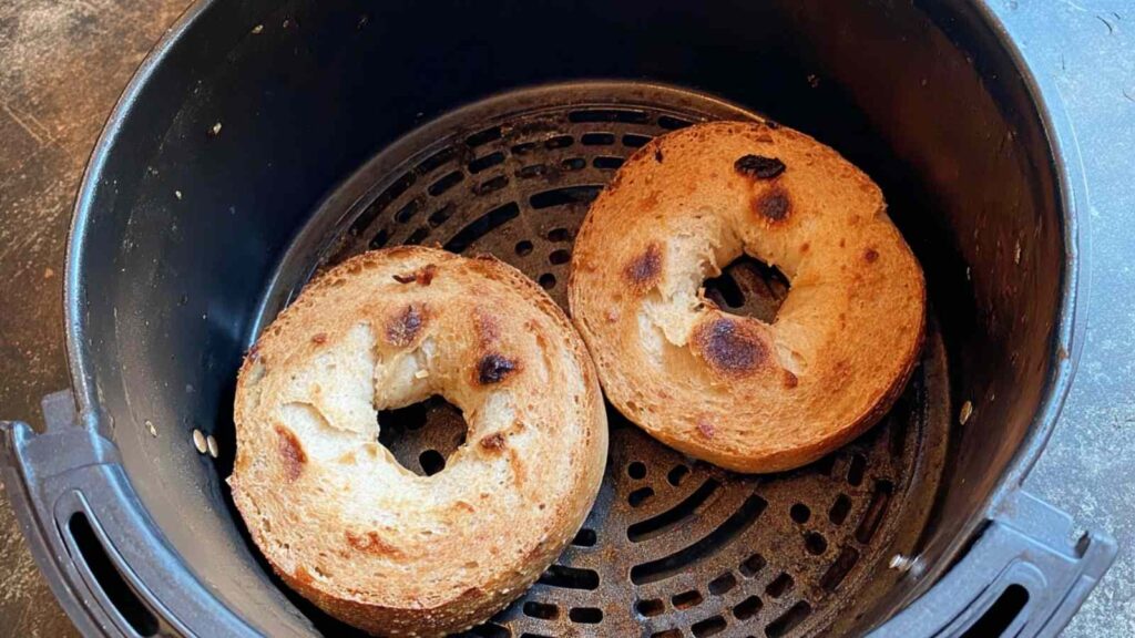 Two bagel halves in an air fryer basket, toasted to a golden brown with visible crispy spots