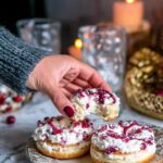 A hand holding a bitten sweet cottage cheese bagel topped with whipped cream and berry preserve, with cozy background.