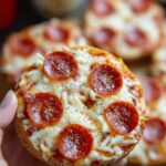 Close-up of a hand holding a pepperoni bagel bite with golden cheese and marinara.