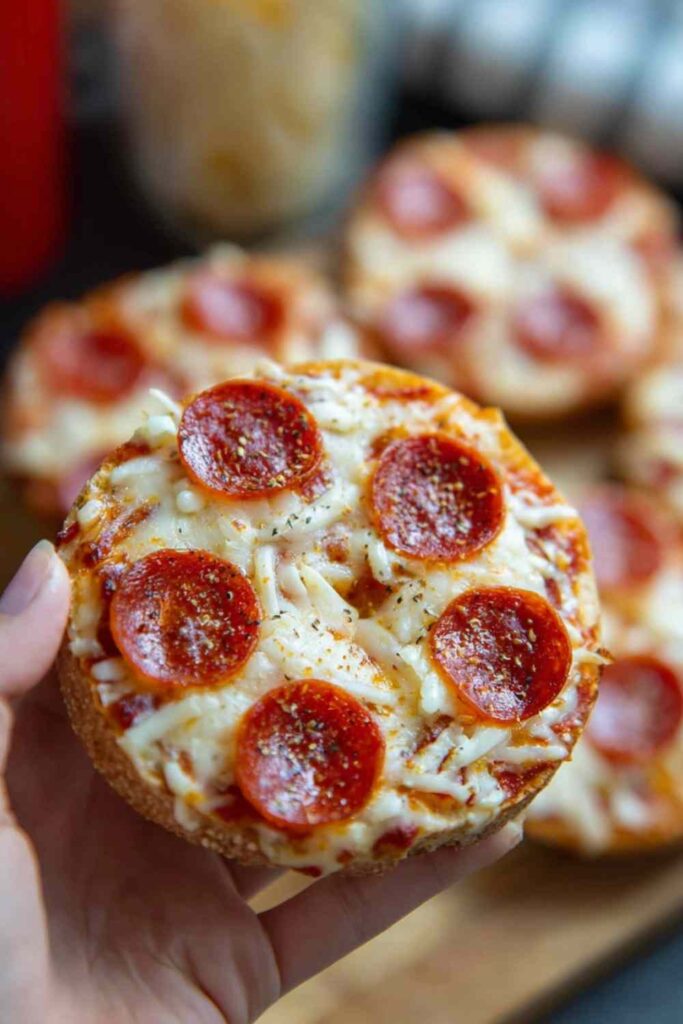 Close-up of a hand holding a pepperoni bagel bite with golden cheese and marinara.