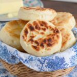 Stacked homemade English muffins in a wicker basket lined with a blue cloth, with a plate of butter in the background