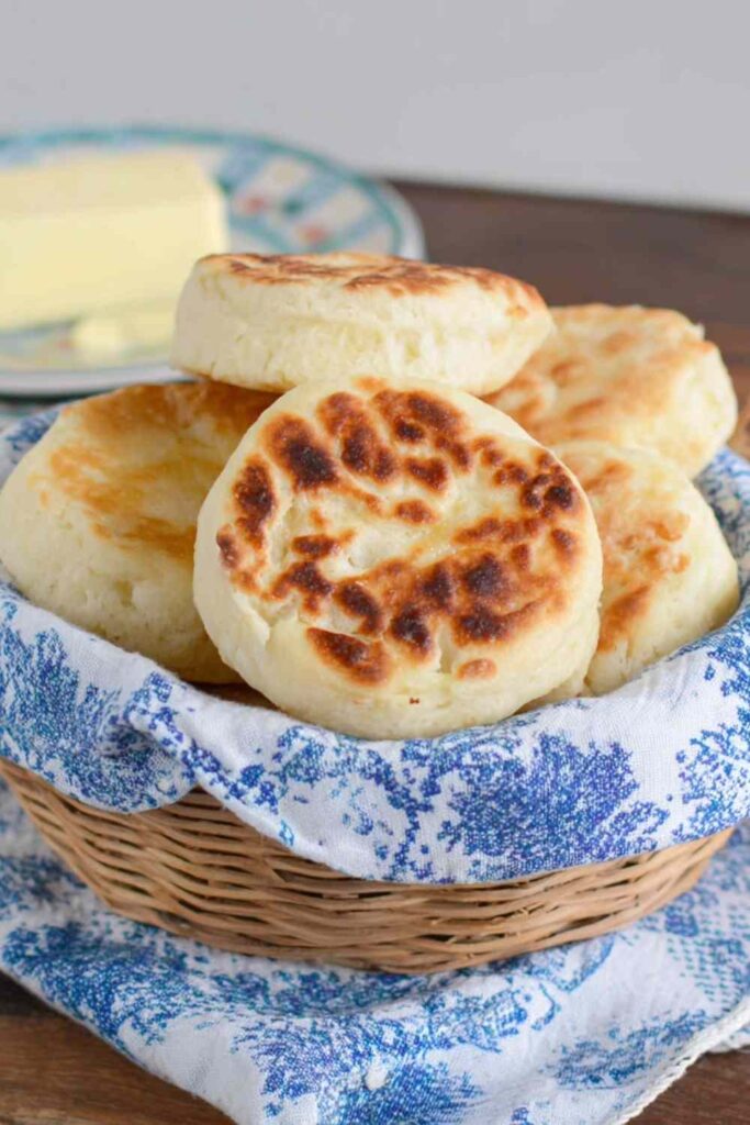 Stacked homemade English muffins in a wicker basket lined with a blue cloth, with a plate of butter in the background