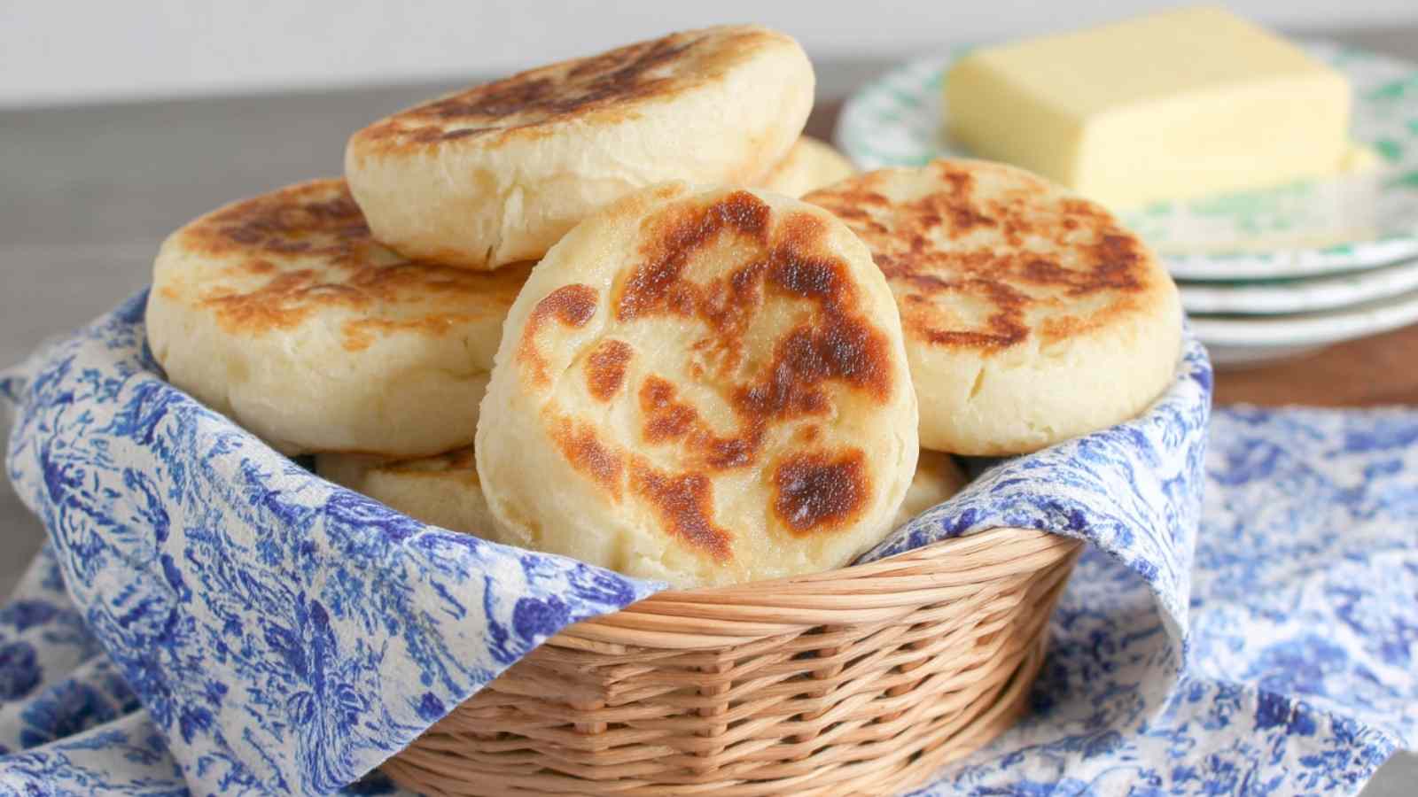 A basket of golden homemade English muffins with crisp tops, nestled in a blue-patterned cloth, with butter in the background
