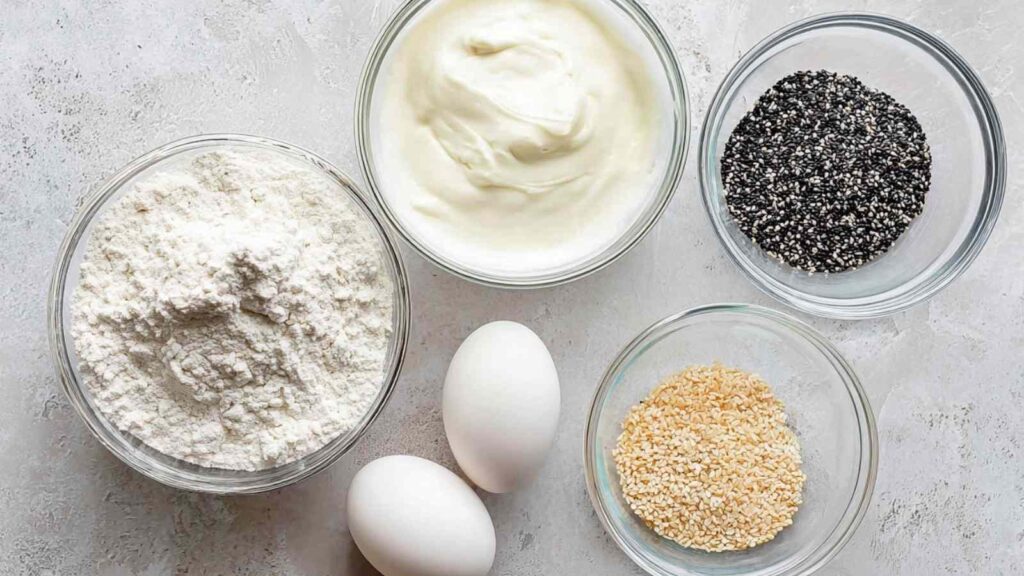Bowls filled with flour, Skyr yogurt, sesame seeds, poppy seeds, and eggs on a white background