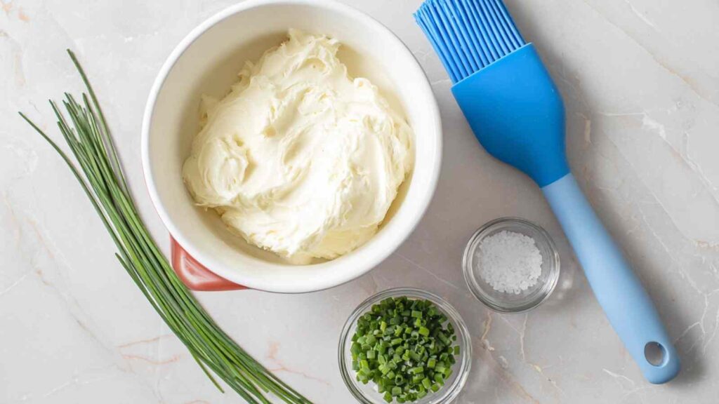 Ingredients laid out for chive cream cheese including cream cheese block, chopped chives, sea salt, and a blue silicone brush
