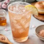Glass of iced Japanese pink salt drink garnished with lemon slice, surrounded by Himalayan salt, honey jar, and fresh lemons
