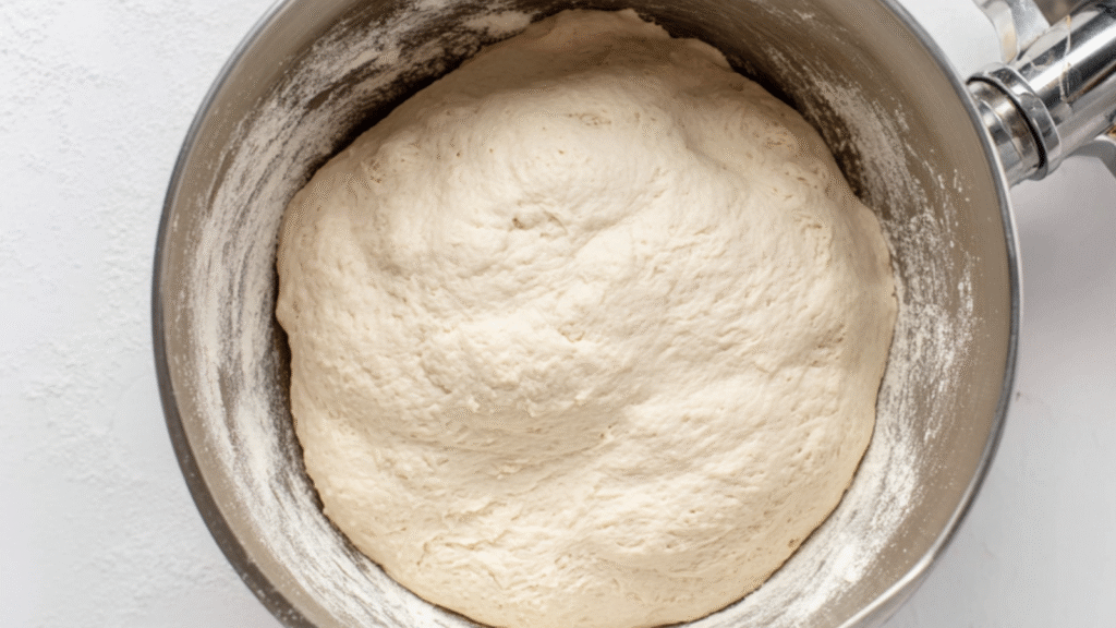 Smooth, risen bagel dough in a metal bowl after kneading, showing a light, tacky surface