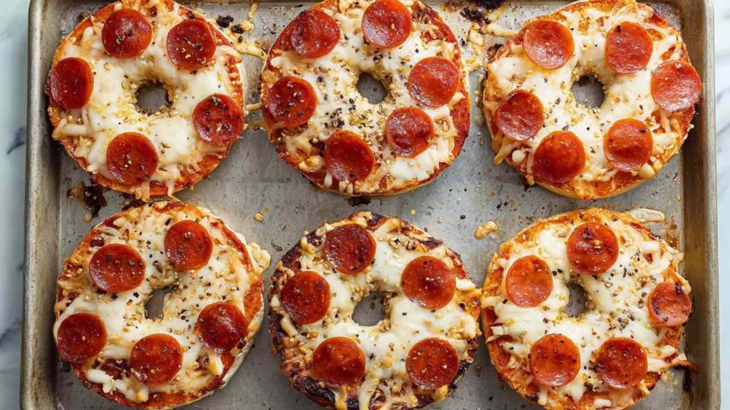 Mini pizza bagels in progress on a baking tray—some with sauce, some with cheese, and others topped with pepperoni.