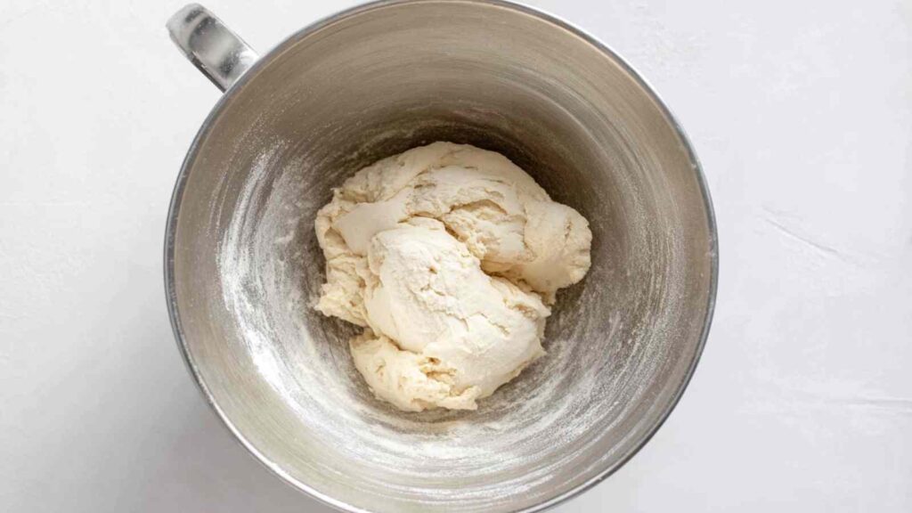 Freshly mixed mini bagel dough in a large stainless steel mixing bowl dusted with flour.