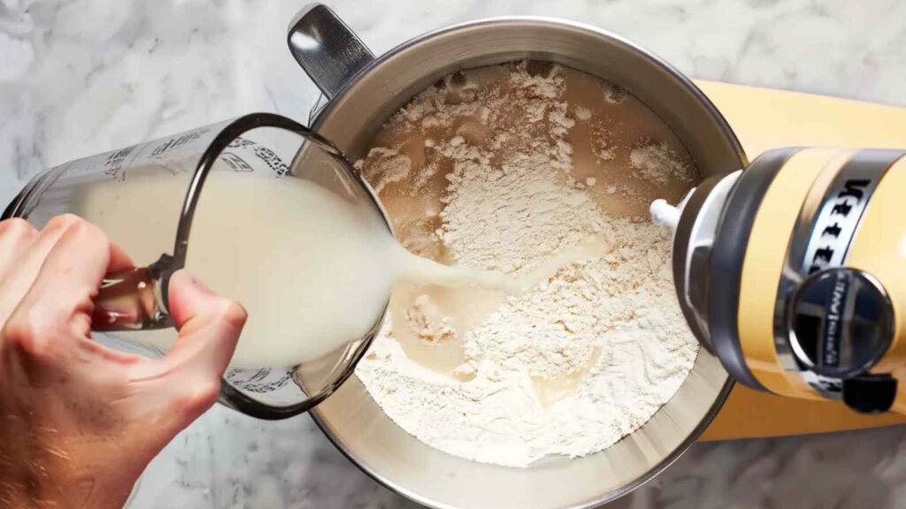 Hand pouring water into a mixing bowl of flour with a stand mixer beside