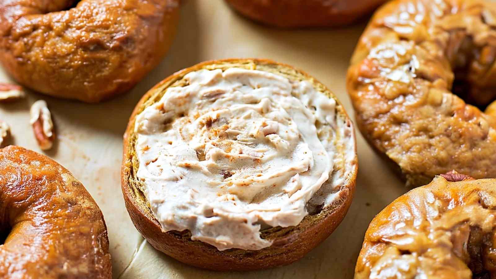 Sliced pumpkin bagel topped with cinnamon-spiced cream cheese, served on parchment with whole pecans nearby.