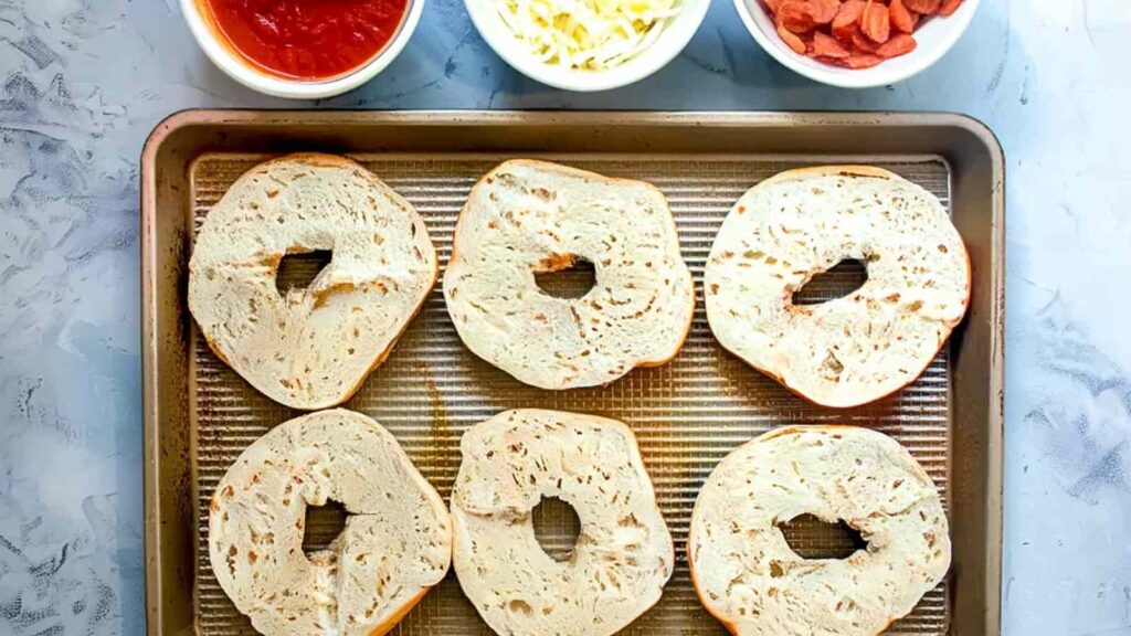 Mini pizza bagels topped with melted cheese and pepperoni served on a white plate, tray of bagels blurred in the background.
