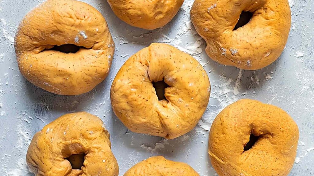 Unbaked pumpkin bagels arranged on a floured surface, showing their round, formed shape.