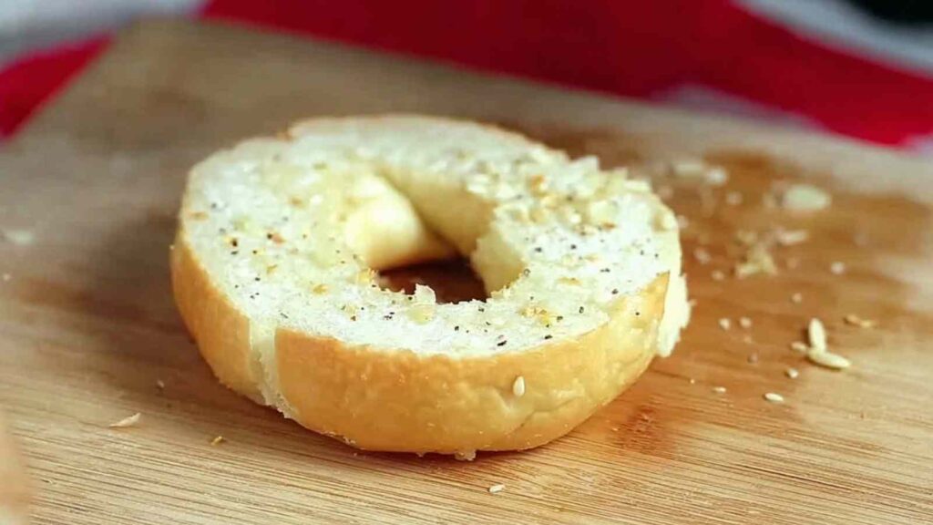 Close-up of a sliced plain bagel showing soft, chewy texture with bits of seasoning on a wooden cutting board.