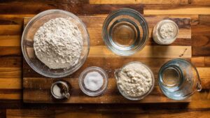 Sourdough bread ingredients arranged on wooden counter including bolted flour, bread flour, active sourdough starter, filtered water, and sea salt in glass bowls and measuring cups