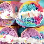 Sliced rainbow bagels stacked and filled with cream cheese and rainbow sprinkles, highlighting vibrant interior swirls.