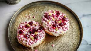 Two sweet cottage cheese bagels topped with white glaze, cherry preserves, crushed candies, and pistachios on a decorative tray.