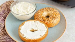 Toasted everything bagel cut in half, one half spread with cream cheese, served on a speckled blue plate with a bowl of cream cheese