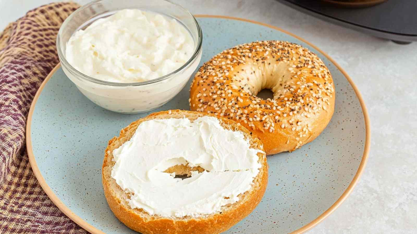 Toasted everything bagel cut in half, one half spread with cream cheese, served on a speckled blue plate with a bowl of cream cheese