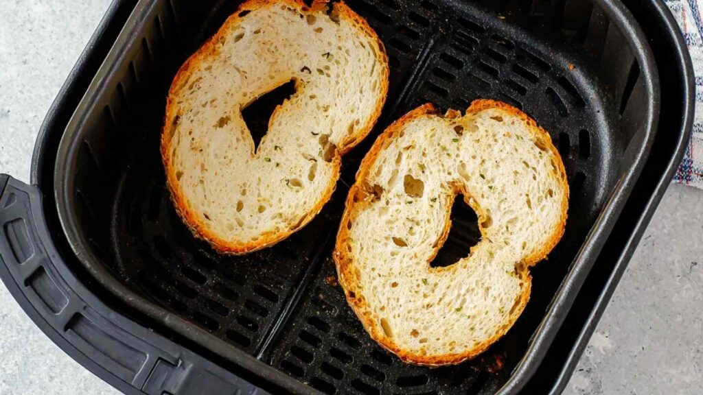 Two plain bagel halves toasting cut side up in an air fryer basket
