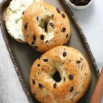 Two vegan sun dried tomato bagels, one sliced and spread with vegan pesto cream cheese on antique tray with a bowl of raisins