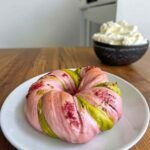Vertical shot of a raspberry pistachio sourdough bagel on a white plate with whipped cream in background