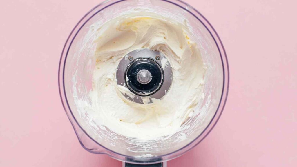 Whipped cream cheese inside a food processor bowl on a pink background.