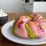Whole raspberry pistachio sourdough bagel on a white plate with pink and green swirls, served next to a bowl of whipped cream