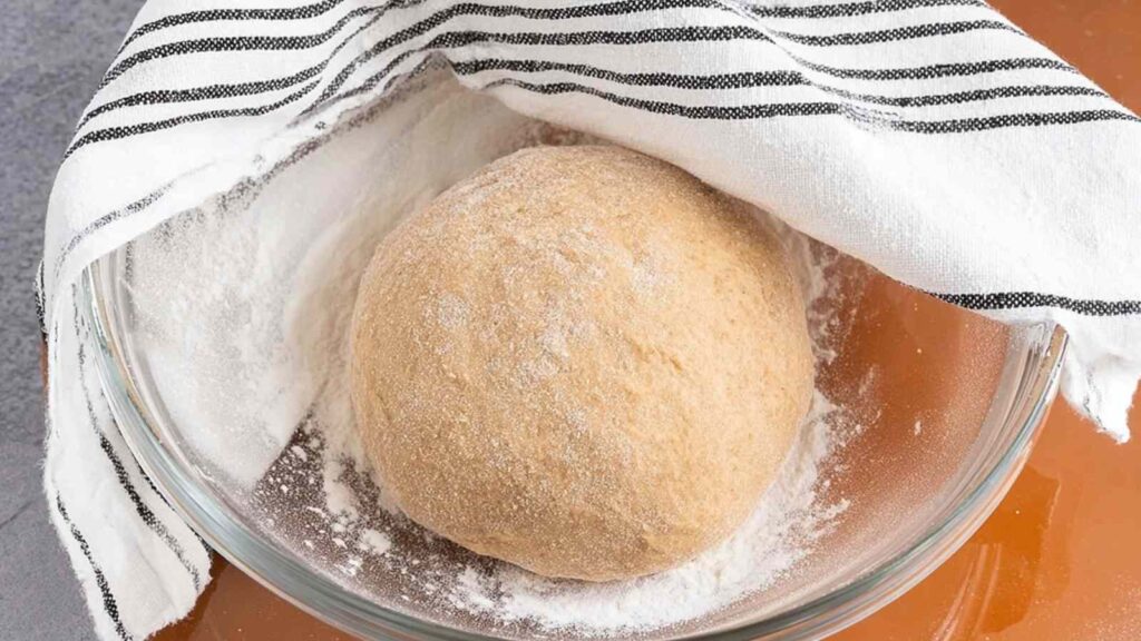 Ball of whole wheat bagel dough resting in a glass bowl covered partially with a kitchen towel