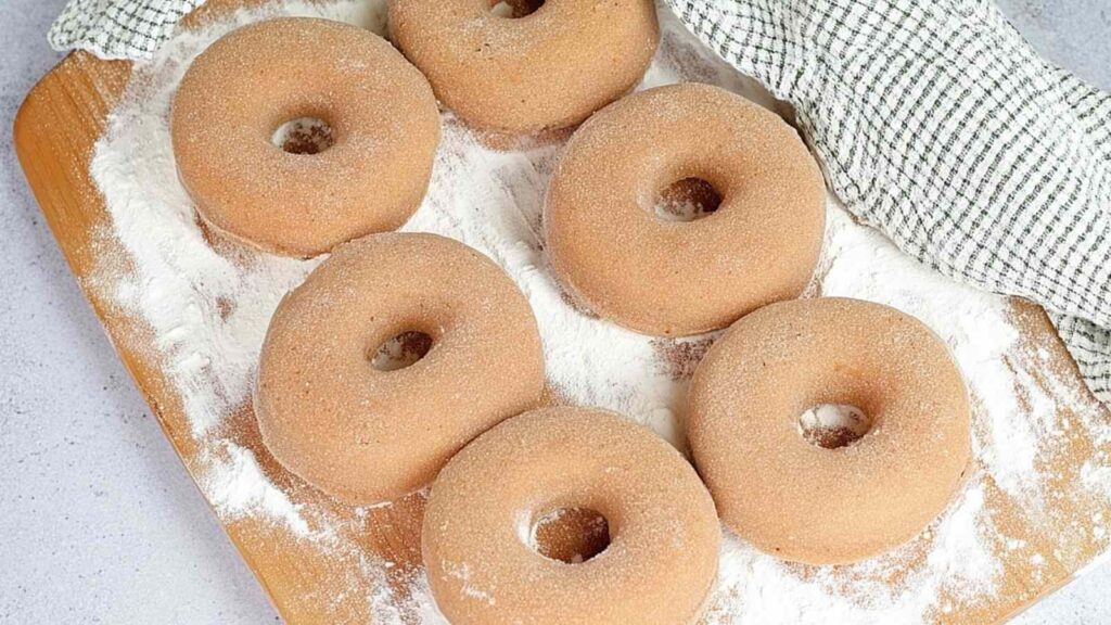 Unbaked whole wheat bagels shaped and resting on a floured wooden surface, covered partially with a towel